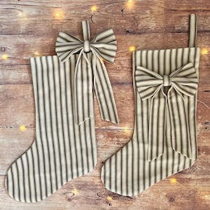 May include: Two Christmas stockings made of beige fabric with vertical dark brown stripes. Each stocking features a matching fabric bow attached to the upper section and a fabric loop at the top for hanging. The design is consistent on both items.