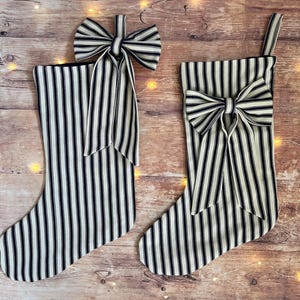 May include: Two festive stockings crafted from cream-coloured fabric featuring thin black vertical stripes. Each stocking is adorned with a large, matching fabric bow on the upper section. One stocking includes a hanging loop, whilst the other has a bow.