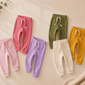 May include: A collection of five pairs of sweatpants in various solid colors including light pink, dark pink, lavender, olive green, cream, and mustard yellow. Each pair features an elastic waistband with a white drawstring and ribbed cuffs.