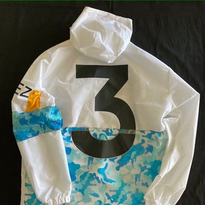 May include: White hooded jacket with a large black number "3" on the back. The jacket has a blue and white camouflage pattern on the bottom half and a yellow and blue stripe on the left sleeve.