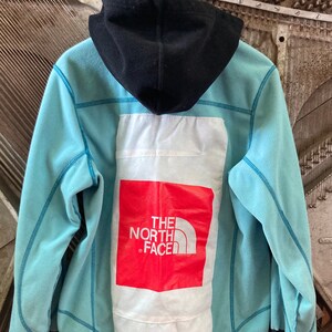 May include: A light blue hooded fleece jacket with a black hood and a white patch on the back with the red logo "THE NORTH FACE" printed on it.