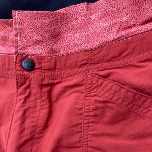 May include: Red pants with a patterned waistband and a button closure. The pants have a pocket on the right side.