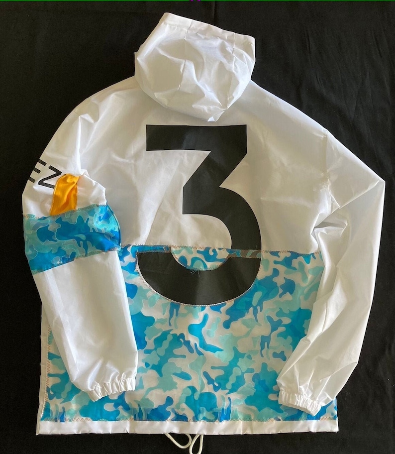 May include: White hooded jacket with a large black number "3" on the back. The jacket has a blue and white camouflage pattern on the bottom half and a yellow and blue stripe on the left sleeve.