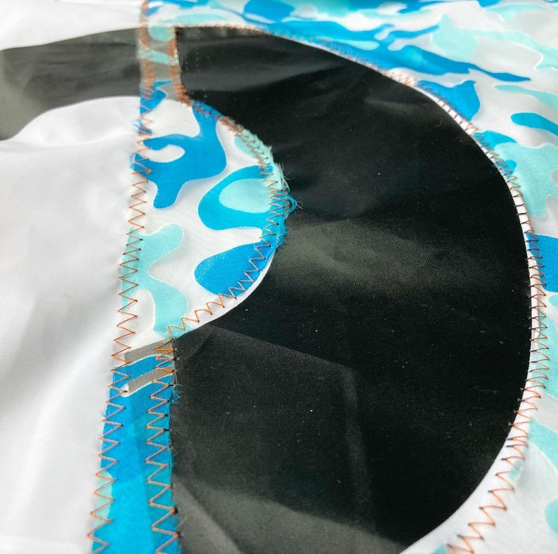 May include: Close-up of a black fabric with a blue and white camouflage pattern sewn around the edges. The fabric is stitched with a copper-colored thread.