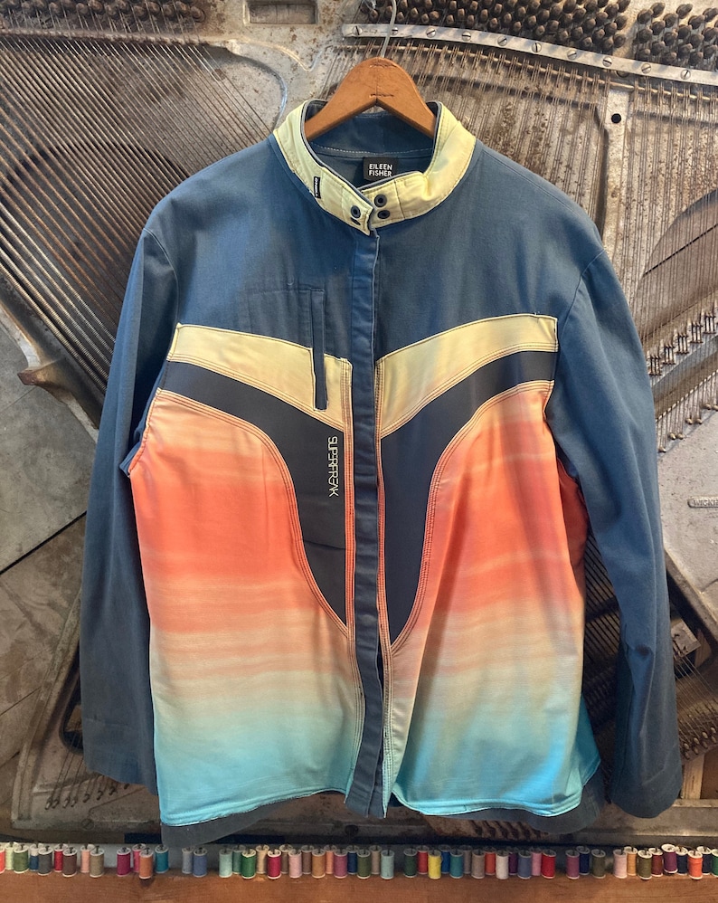 May include: A blue jacket with a color block design. The jacket has a light yellow collar and a front panel with a gradient of orange, pink, and blue. The brand name "EILEEN FISHER" is visible on the collar. The word "SUPERFK" is printed on the front.