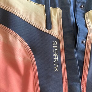 May include: Close-up of a navy blue and peach-colored garment with beige accents. The garment has a stitched design and the word "SUPERFREAK" is printed vertically in gold lettering. The garment has black buttons.