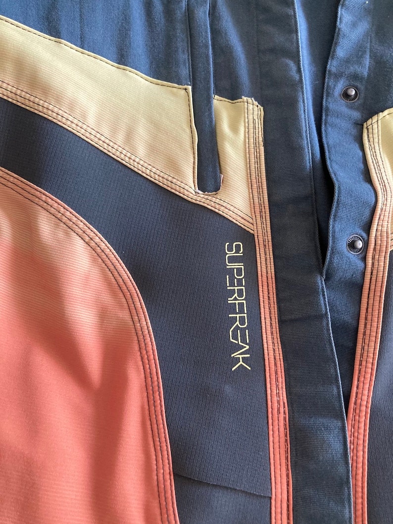 May include: Close-up of a navy blue and peach-colored garment with beige accents. The garment has a stitched design and the word "SUPERFREAK" is printed vertically in gold lettering. The garment has black buttons.