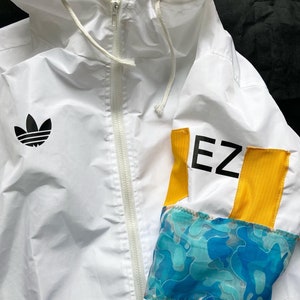 May include: White windbreaker jacket with a black Adidas logo on the left chest and a yellow and blue patterned sleeve with the text "EZ" in black.