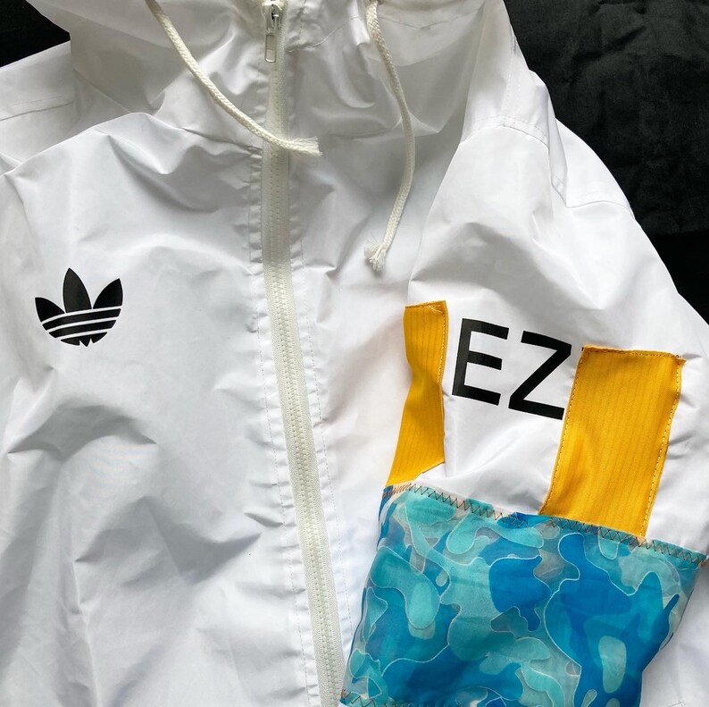 May include: White windbreaker jacket with a black Adidas logo on the left chest and a yellow and blue patterned sleeve with the text "EZ" in black.