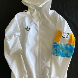 May include: White hooded jacket with a black Adidas logo on the left chest and a blue and yellow camouflage pattern on the right sleeve. The sleeve also has the text "EZ" in black.