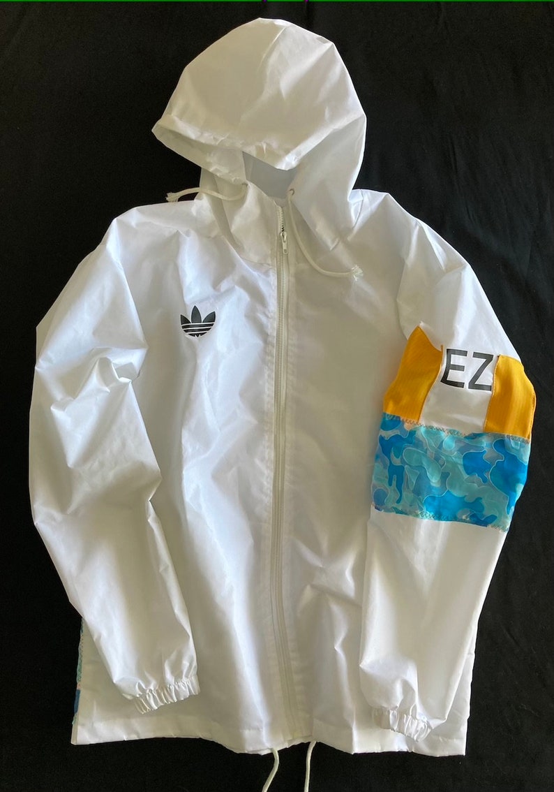 May include: White hooded jacket with a black Adidas logo on the left chest and a blue and yellow camouflage pattern on the right sleeve. The sleeve also has the text "EZ" in black.