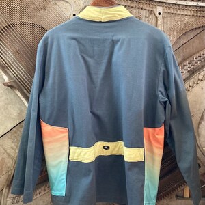 May include: A long-sleeved, blue jacket with a yellow collar and accents. The jacket features a color gradient on the lower pockets, transitioning from orange to turquoise. The back of the jacket is visible.
