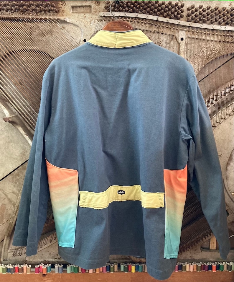 May include: A long-sleeved, blue jacket with a yellow collar and accents. The jacket features a color gradient on the lower pockets, transitioning from orange to turquoise. The back of the jacket is visible.