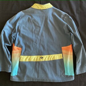 May include: Blue jacket with a yellow collar and accents. The jacket features a horizontal yellow band across the back and colorful gradient panels in shades of orange, yellow, and blue. The jacket has long sleeves.