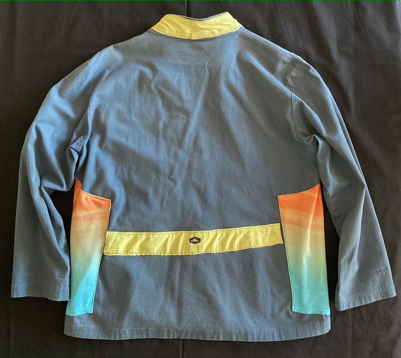 May include: Blue jacket with a yellow collar and accents. The jacket features a horizontal yellow band across the back and colorful gradient panels in shades of orange, yellow, and blue. The jacket has long sleeves.