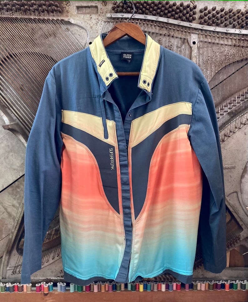May include: A blue jacket with a color gradient design in orange, yellow, and blue. The jacket has a light yellow collar and accents, with the text "SUPERFREAK" on the front. The brand name "EILEEN FISHER" is visible on the collar.