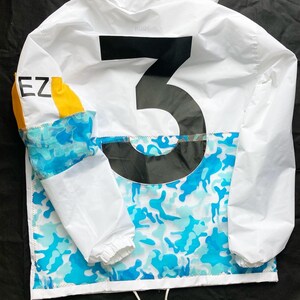 May include: White jacket with a large black number "3" on the back. The jacket has a blue and white camouflage pattern on the bottom and a yellow and blue panel on the left sleeve. The text "EZ" is visible on the sleeve.