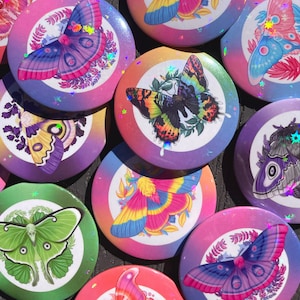 May include: A collection of 10 round, colourful, iridescent buttons featuring various butterfly and moth designs. Each button has a different colour gradient background and a different insect design. The buttons are perfect for adding a touch of whimsy to any project.