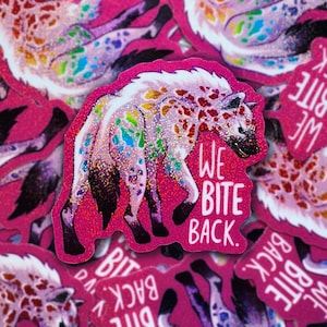 Könnte beinhalten: Ein pinkfarbener Glitzeraufkleber mit einer Illustration einer gefleckten Hyäne mit Regenbogenakzenten. Der Text "WE BITE BACK" ist in Weiß geschrieben. Ein lustiges, auffälliges Design.