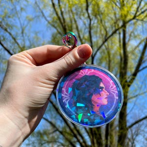 May include: A round, iridescent, holographic charm featuring a portrait of a person with dark hair and a pink background. The charm is attached to a silver heart-shaped ring.