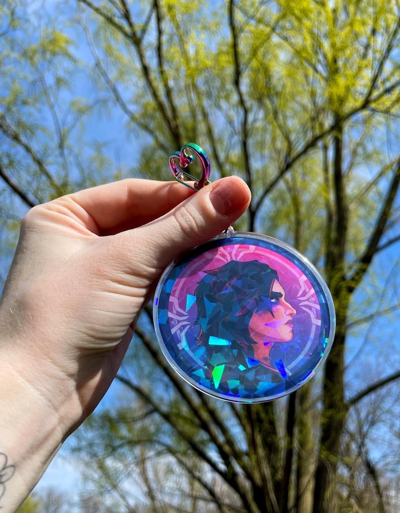 May include: A round, iridescent, holographic charm featuring a portrait of a person with dark hair and a pink background. The charm is attached to a silver heart-shaped ring.