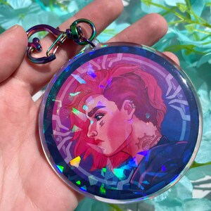 May include: A round, iridescent keychain featuring a colorful illustration of a person with pink hair and a tattoo. The keychain has a silver metal clasp.