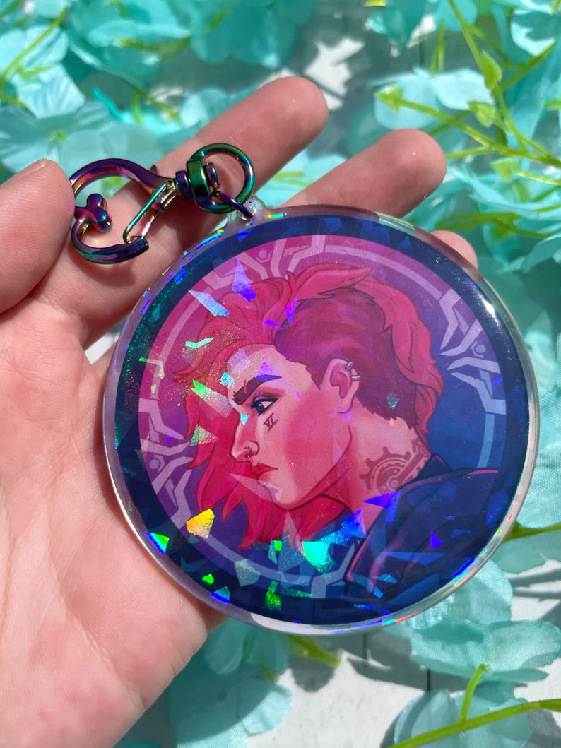 May include: A round, iridescent keychain featuring a colorful illustration of a person with pink hair and a tattoo. The keychain has a silver metal clasp.