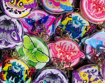May include: Assortment of colorful, iridescent keychains featuring various moth and butterfly designs. Each keychain has a clear, round, plastic casing with a silver star charm.
