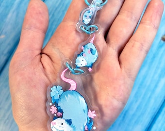 May include: A clear acrylic keychain featuring three connected charms of opossums in varying sizes. The charms display blue and white opossums with pink tails and feet, accented by small blue and pink flowers. A light blue lobster clasp is attached.