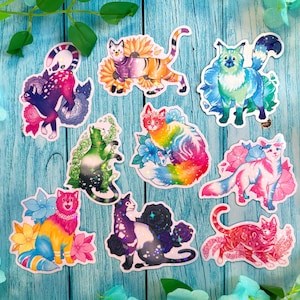 May include: A collection of colorful cat stickers. Each sticker features a unique cat design with vibrant colors and floral accents. The cats are depicted in various poses, with a whimsical and artistic style. The stickers are on a blue wooden background.