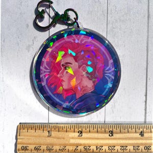 May include: A holographic round keychain featuring a colorful illustration of a person with pink hair and a blue and purple background. The keychain has a silver heart-shaped ring.