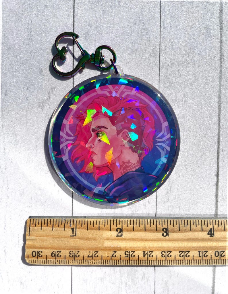May include: A holographic round keychain featuring a colorful illustration of a person with pink hair and a blue and purple background. The keychain has a silver heart-shaped ring.