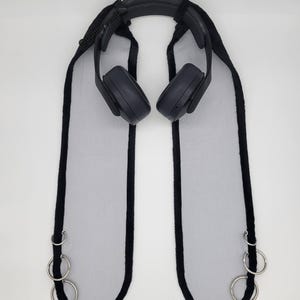 May include: Black over-ear headphones with a padded headband and earcups. The headphones are attached to a black and white accessory with three silver rings on each side. The accessory has a black trim.