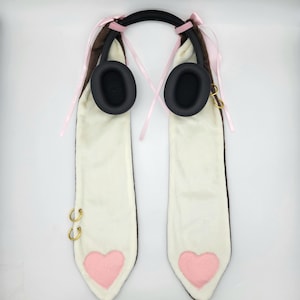 May include: Headphones with a black headband and earcups, featuring long, white faux fur ear covers with brown trim. Pink heart-shaped accents and gold-coloured hoops adorn the bottom of the ear covers. Pink ribbons are tied to the headband.