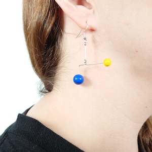 Puede incluir: Un par de pendientes de plata con un diseño geométrico azul y amarillo. Los pendientes están hechos de alambres metálicos finos y tienen dos cuentas redondas, una azul y una amarilla.