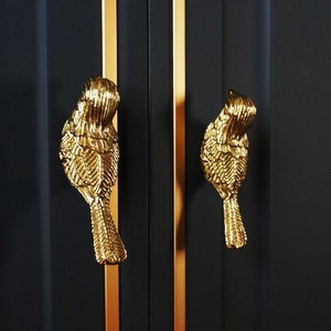 Puede incluir: Dos pomos de armario dorados en forma de pájaro con diseños detallados de plumas sobre un fondo negro con ribetes dorados.