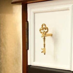 Puede incluir: Un tirador de cajón en forma de llave dorada con detalles ornamentados, montado en un frente de cajón de madera pintado de blanco.
