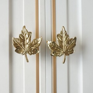 Puede incluir: Dos tiradores de puerta de armario en forma de hoja de arce de color dorado. Las hojas son detalladas y tienen un acabado ligeramente texturizado.