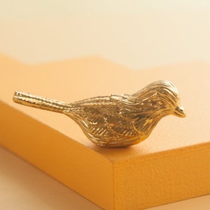 May include: A small, gold-colored bird figurine with detailed feathers. The bird is perched on a light orange surface.