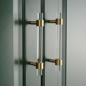 Pode incluir: Duas puxadores de armário de acrílico transparente com ferragens de cor latão. Os puxadores cilíndricos são montados em uma superfície verde escuro. As ferragens de latão conectam as hastes de acrílico transparente ao armário.