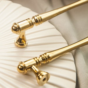 Peut inclure: Deux poignées de porte de placard dorées avec un design décoratif cannelé. Les poignées ont une extrémité arrondie en forme de bouton et une longue poignée fine.