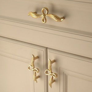 Op de afbeelding: Gouden kleurige kastbeslag met een decoratief strikdesign. Het beslag heeft de vorm van een strik met een lus aan de bovenkant. Het beslag is gemonteerd op een witte kastdeur.