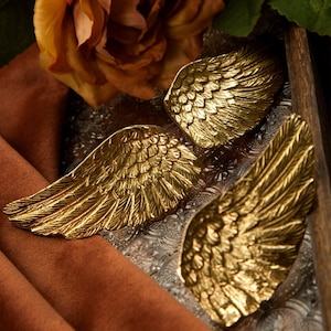 May include: Two gold metal angel wing napkin rings. The wings are detailed with a textured pattern.