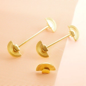 Op de afbeelding: Drie gouden kastknoppen met een waaiervormig ontwerp. De knoppen bevinden zich op een roze achtergrond.