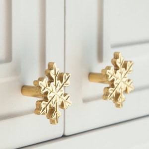 Op de afbeelding: Twee gouden kastknoppen in de vorm van sneeuwvlokken met een gedetailleerd ontwerp. De knoppen zijn gemonteerd op een witte kastdeur.