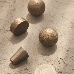 May include: Five hammered brass cabinet knobs in different shapes, including round, oval, and cone. The knobs have a rustic, antique finish.