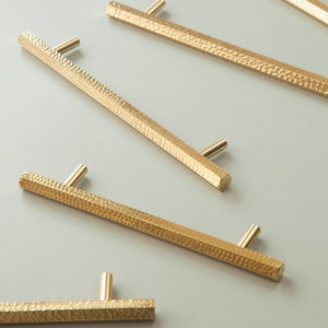 May include: Five gold-toned cabinet pulls with a hammered texture. The pulls are rectangular and have a slightly curved shape.