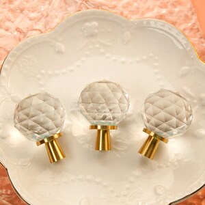 Puede incluir: Tres pomos de armario de cristal transparente con bases de metal dorado. Los pomos son facetados y tienen forma redonda.