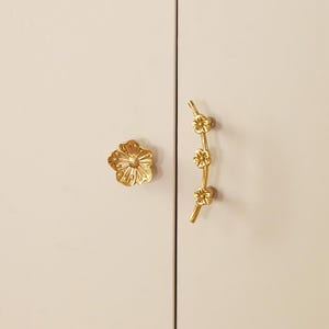 Puede incluir: Herrajes dorados para armarios, que incluyen un pomo en forma de flor y un tirador en forma de rama con tres acentos florales. Los herrajes están montados en una puerta de armario de color claro.