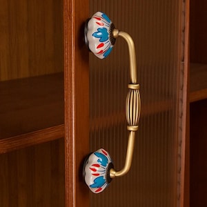 May include: Cabinet handle with a floral design. The handle features a white ceramic knob with blue and red floral accents. The curved handle is made of brass and is attached to a wooden cabinet door.